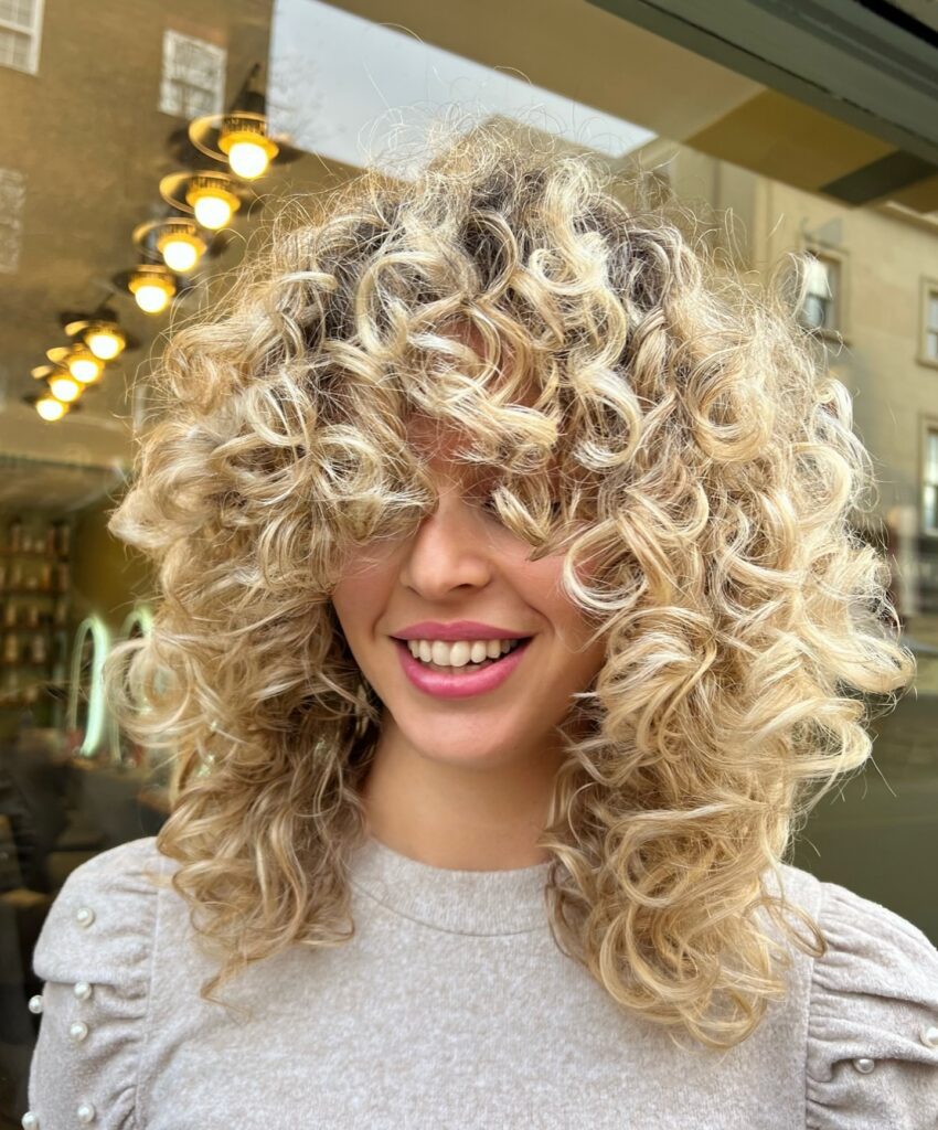 Woman with curly, layered hair and bangs, showcasing retro-inspired 90s haircut trends. Credits: @hairspringstudio