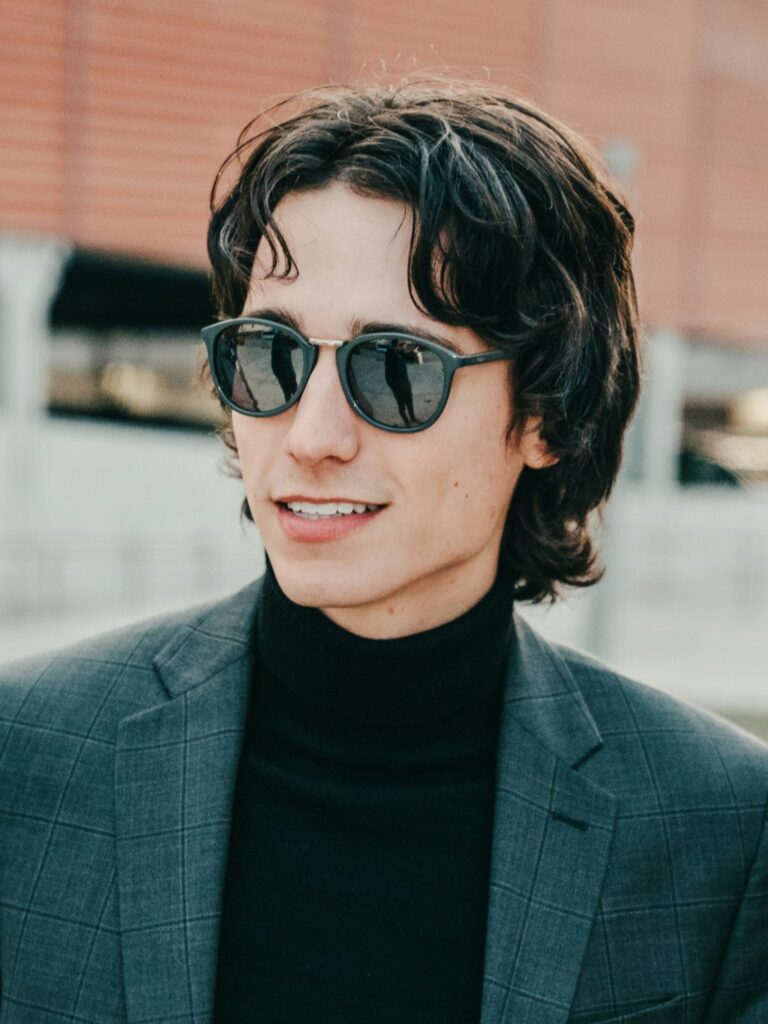 [ALT_TEXT: A young man with a retro mullet haircut wearing a grey suit jacket and sunglasses standing outside smiling.]