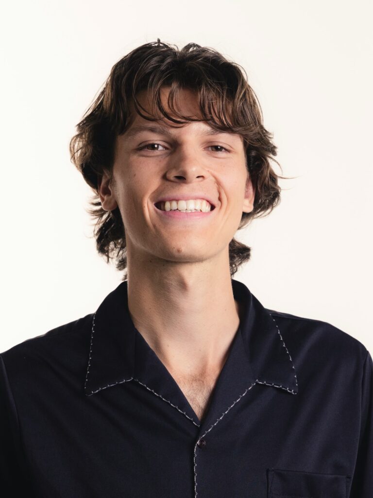 [ALT_TEXT: A young man with a layered, textured mullet haircut wearing a dark blue shirt smiling.]