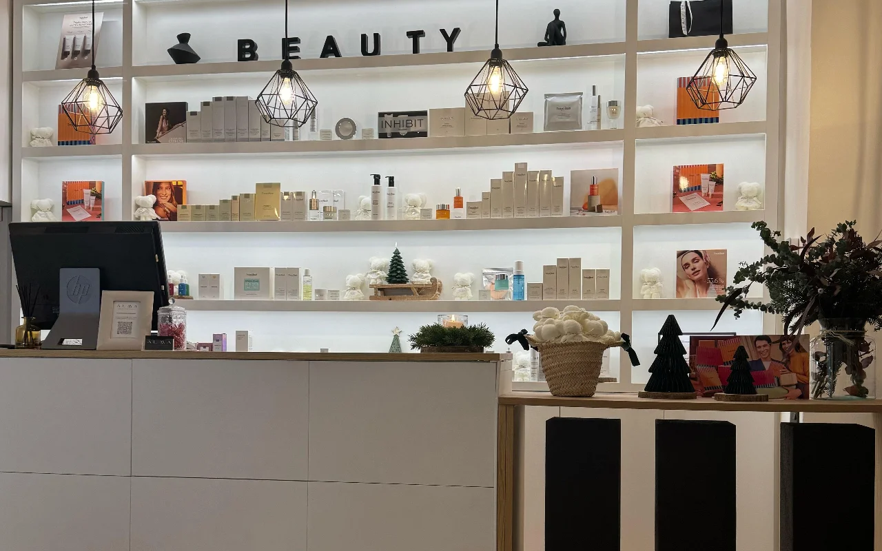 A bright, modern reception area featuring a large, white back-lit shelving unit filled with various skincare products. Geometric black wire pendant lights hang from the ceiling, and the front desk is a clean, white minimalist counter.