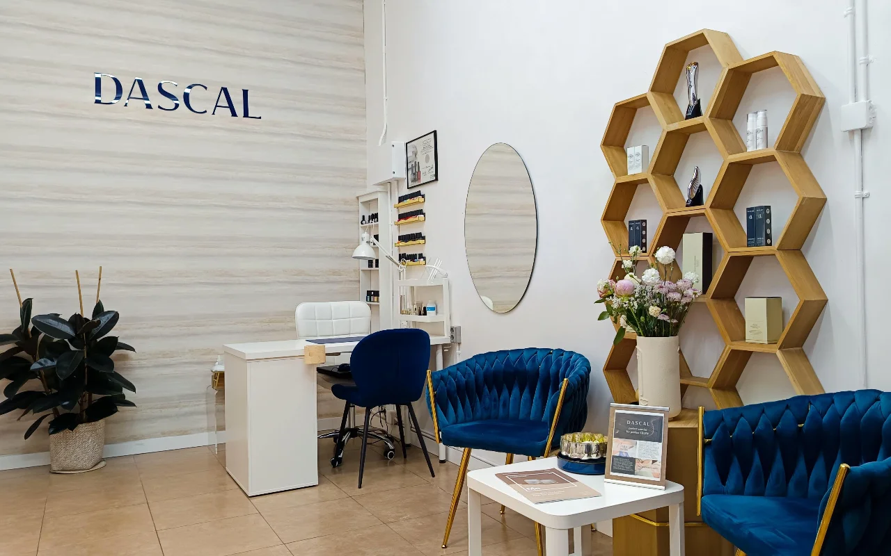A stylish beauty salon interior featuring deep blue velvet chairs and a unique hexagonal wooden shelving unit on the wall. A white manicure desk sits in the corner against a textured, light-colored wall with the word "DASCAL" in blue lettering.