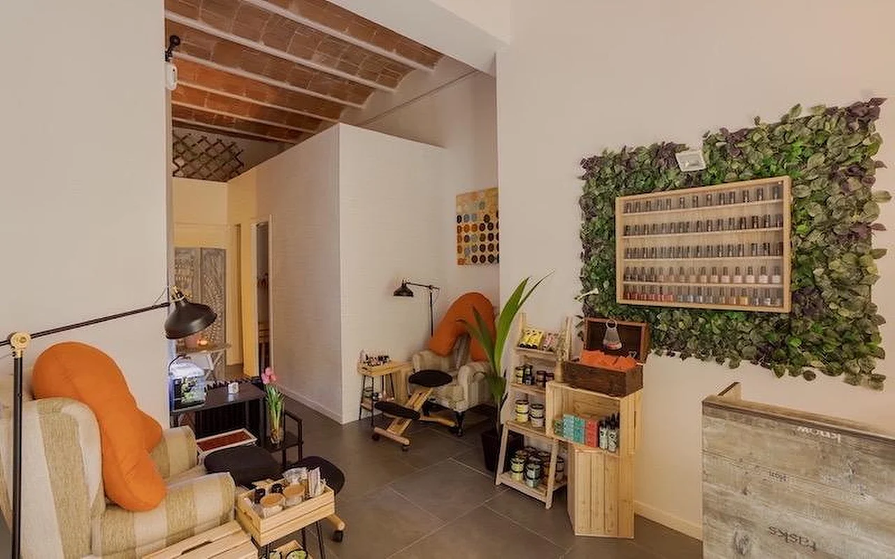 A cozy, bohemian-style nail salon with plush armchairs and orange cushions. The wall features a large vertical plant installation (greenery wall) surrounding a wooden display case filled with nail polish bottles.