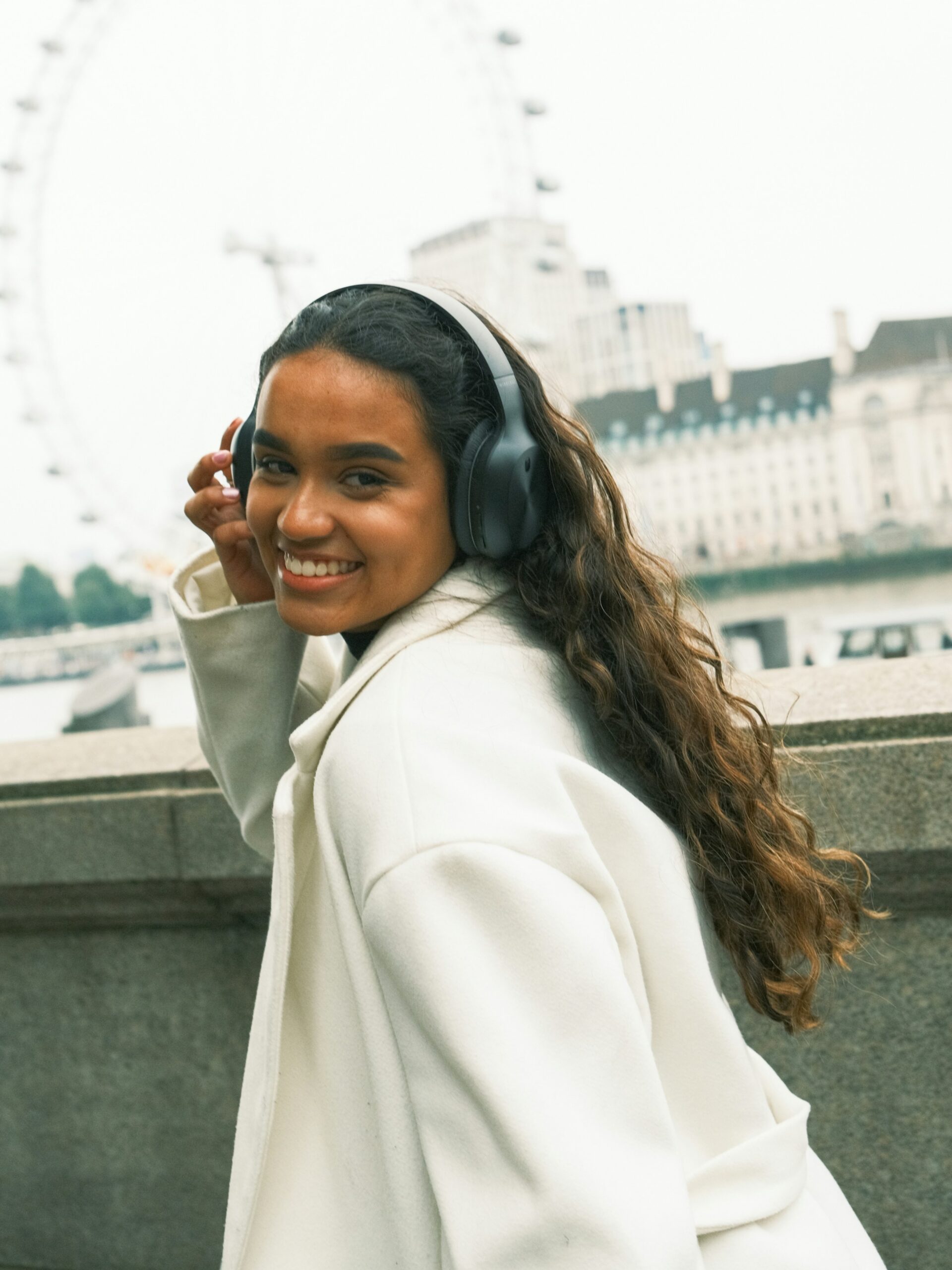 A smiling woman with microblading on her brows wearing black over-ear headphones and a white coat walking through the streets of Brussels
