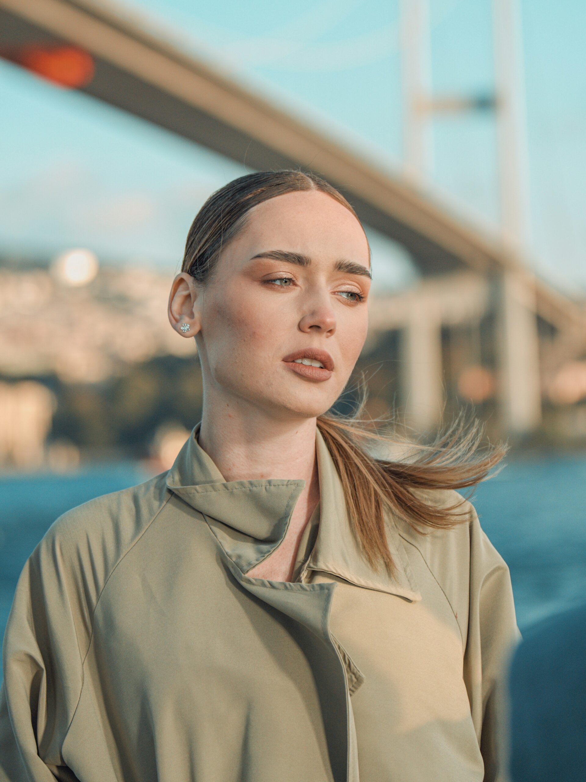 A woman with microblading on her brows and fair hair tied back wearing a trench coat and standing in front of a bridge in Istanbul