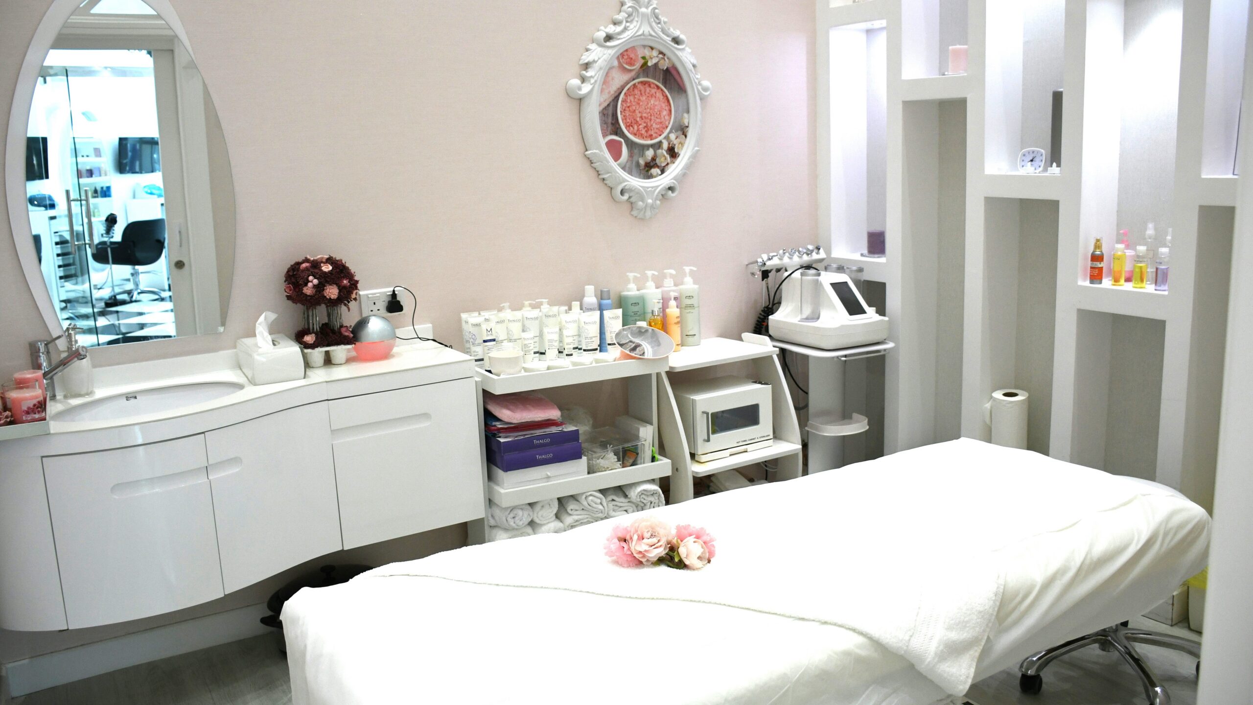 Bright, clean and welcoming salon treatment room with a bed, products on shelves, an ornate mirror and flowers