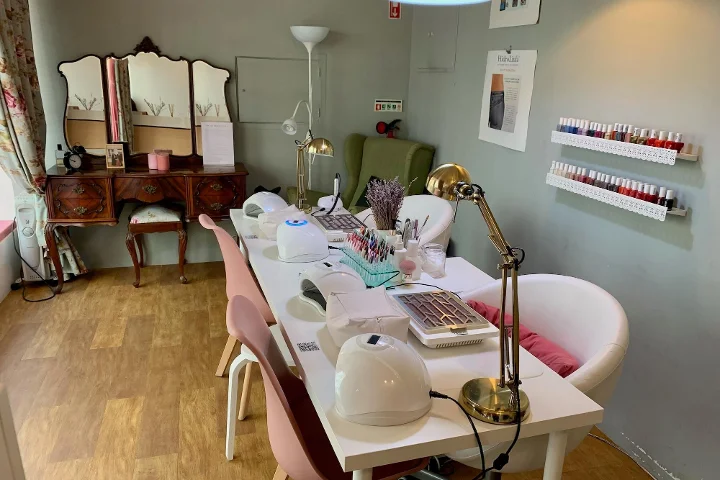 An indoor view of a cosy beauty salon specializing in manicures. In the foreground, a long white table is set up with nail lamps, desk lights, and various nail technician tools, flanked by pink and white chairs. In the background, there is a vintage wooden dressing table with a three-way mirror, a green armchair, and white shelves on the wall displaying rows of colourful nail polish bottles.