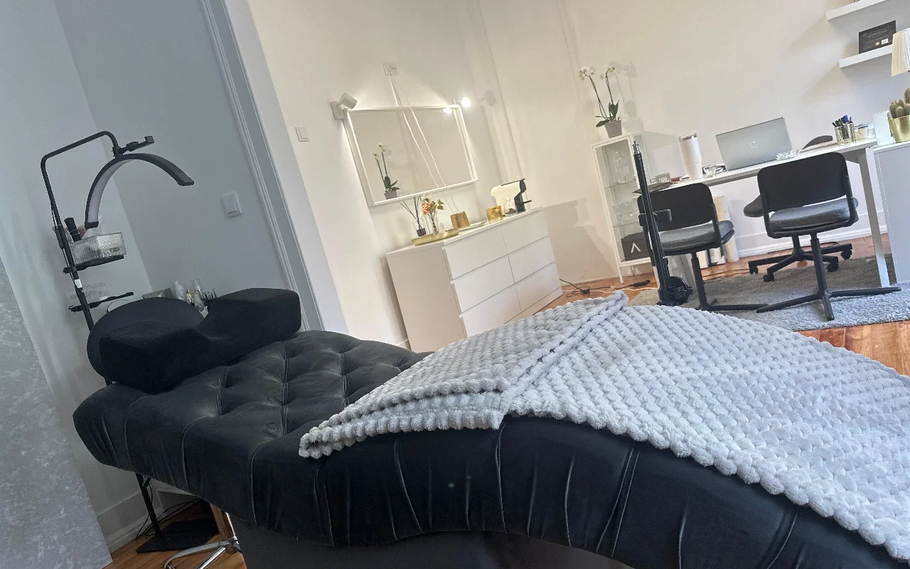 A modern eyelash treatment room captured from a low angle. In the foreground, a plush, black tufted beauty couch is draped with a textured grey blanket and fitted with an ergonomic neck pillow under a curved ring light. The background shows a minimalist white office setup with a desk, two black chairs, a laptop, a white chest of drawers, and a large mirror.
