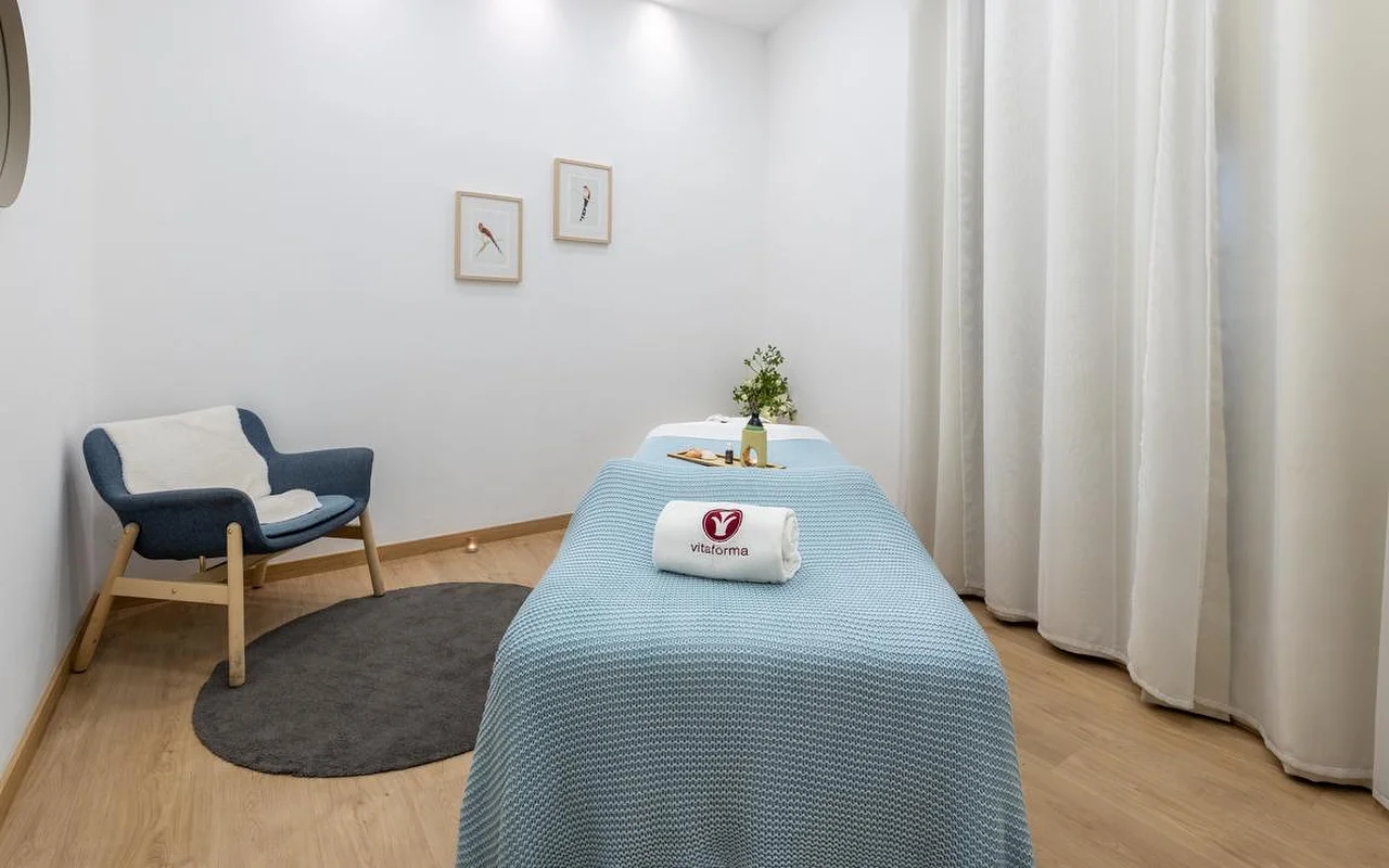 A clean, minimalist spa room with white walls and light wood flooring. A treatment table is neatly made up with a light blue knitted blanket and a rolled white towel bearing the 'Vitaforma' logo. In the corner, a stylish blue armchair sits on a small round grey rug, and long white curtains hang by the window.