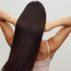 Back view of a woman wearing a white strappy top with her arms up running her fingers through her long, glossy brunette hair
