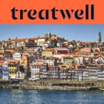 The Ribeira waterfront in Porto, with rows of colourful houses and terracotta rooftops rising up the hillside from the Douro river and the Clérigos tower on the skyline under a clear blue sky.
