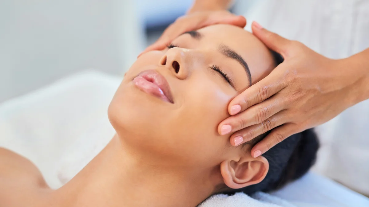 A woman lays down in a beauty salon while a facialist gently holds her hands on her head during a facial