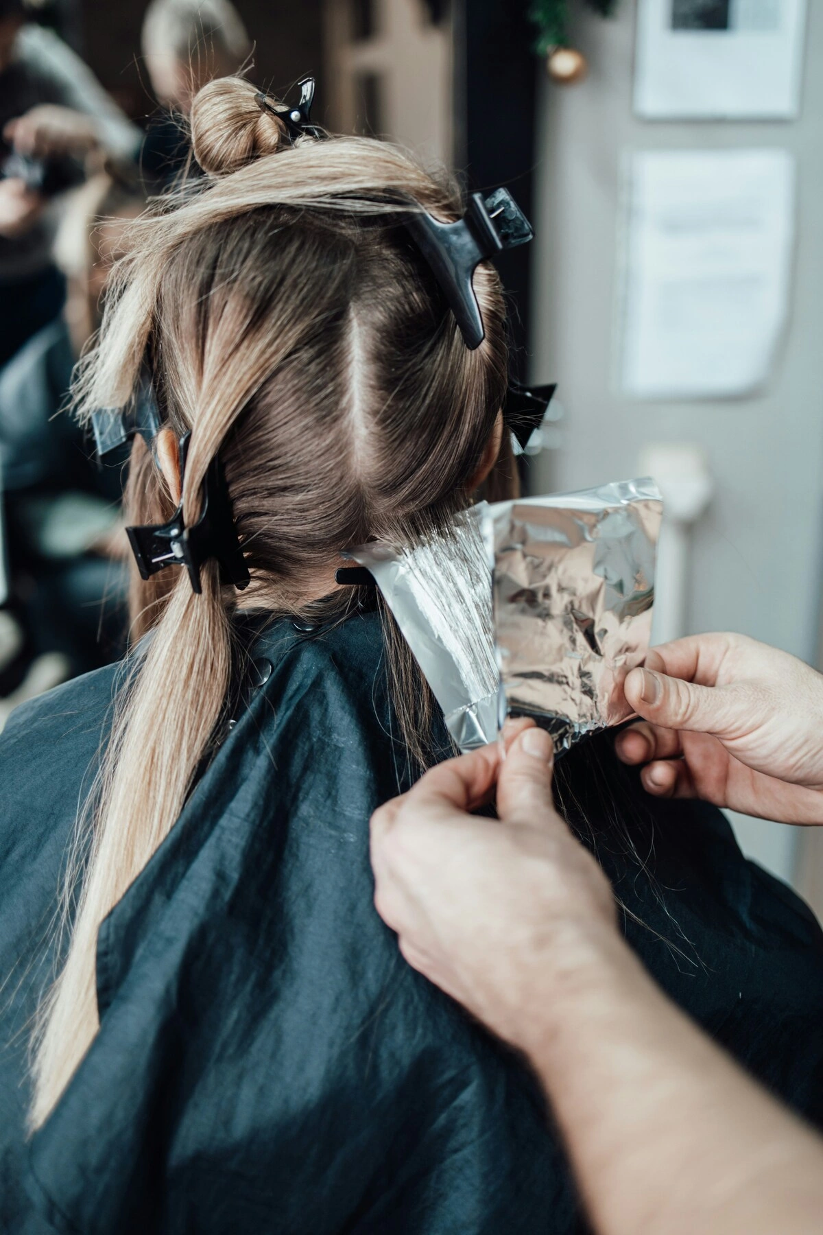 Back view of a client sitting in a chair while a hairdresser folds a foil in her hair during a treatment to apply highlights