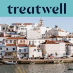 A whitewashed Algarve fishing village of terracotta-roofed houses rising up a hillside above a calm waterway, with small fishing boats moored along the quay, palm trees and a church tower under a clear blue sky.