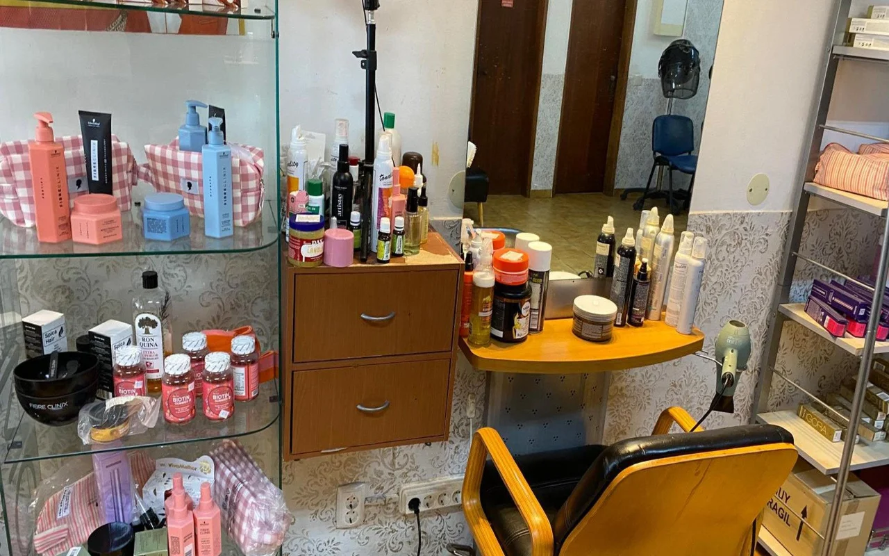 A hairdresser's station with a wall mirror and a wooden counter crowded with styling products, a glass cabinet of hair-care products and supplements alongside, and a classic black-and-wood styling chair.