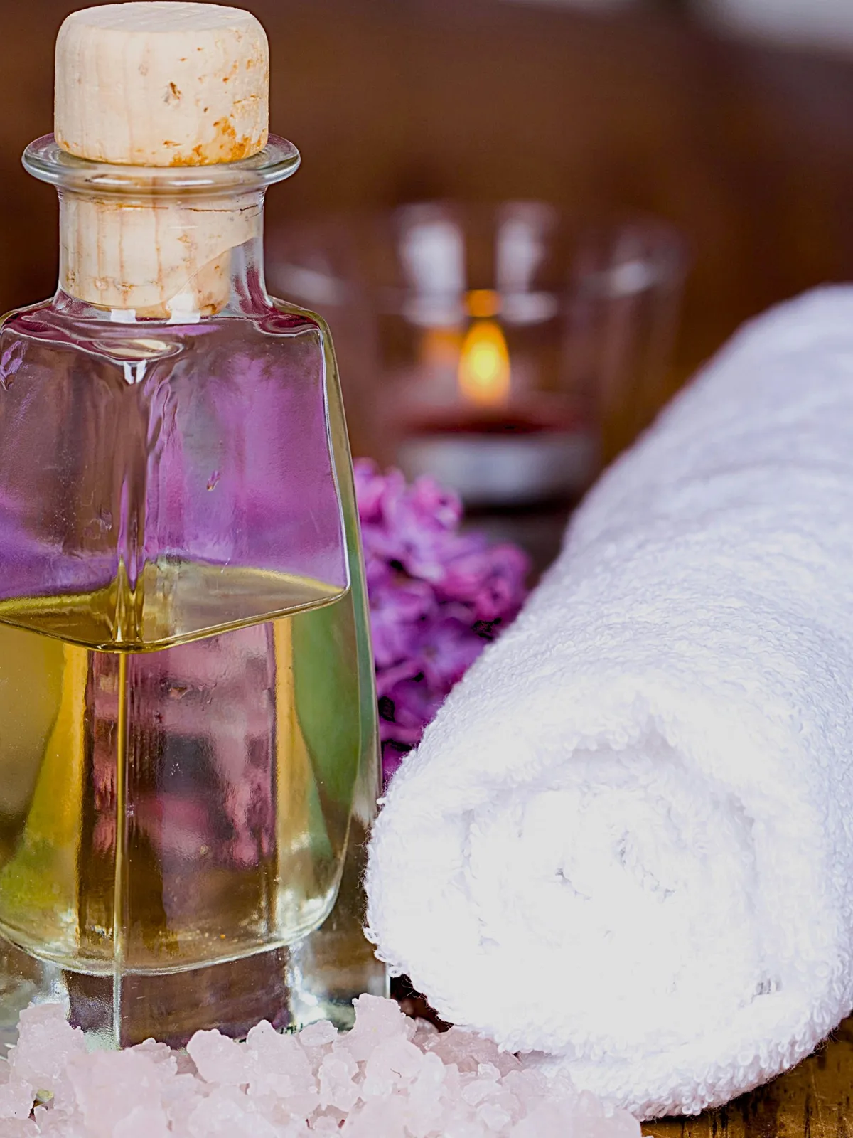 Close-up of a glass bottle of oil with a cork on a table next to a rolled white towel and pink flowers