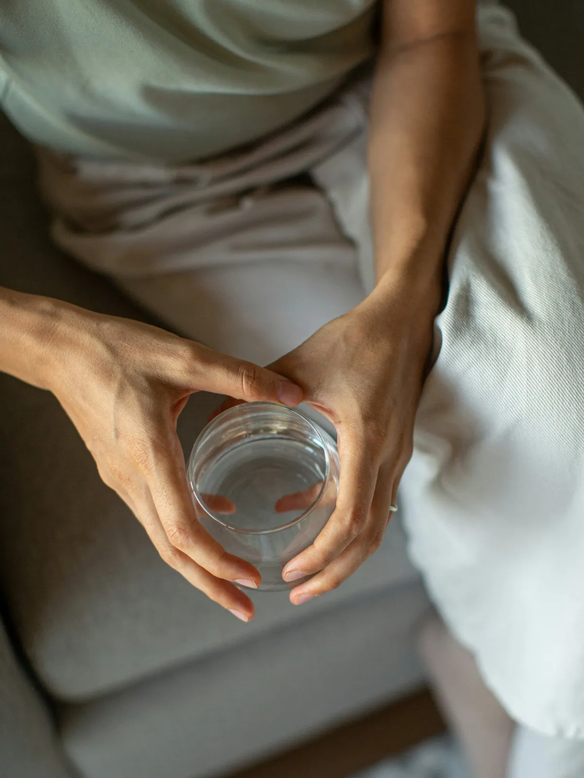 Top view of a person sitting on a grey sofa in loose clothing and holding a glass of water with both hands